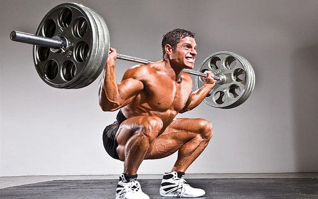 Muscular man performing a barbell back squat with full range of motion, demonstrating proper squat depth and joint mobility for maximum muscle activation.